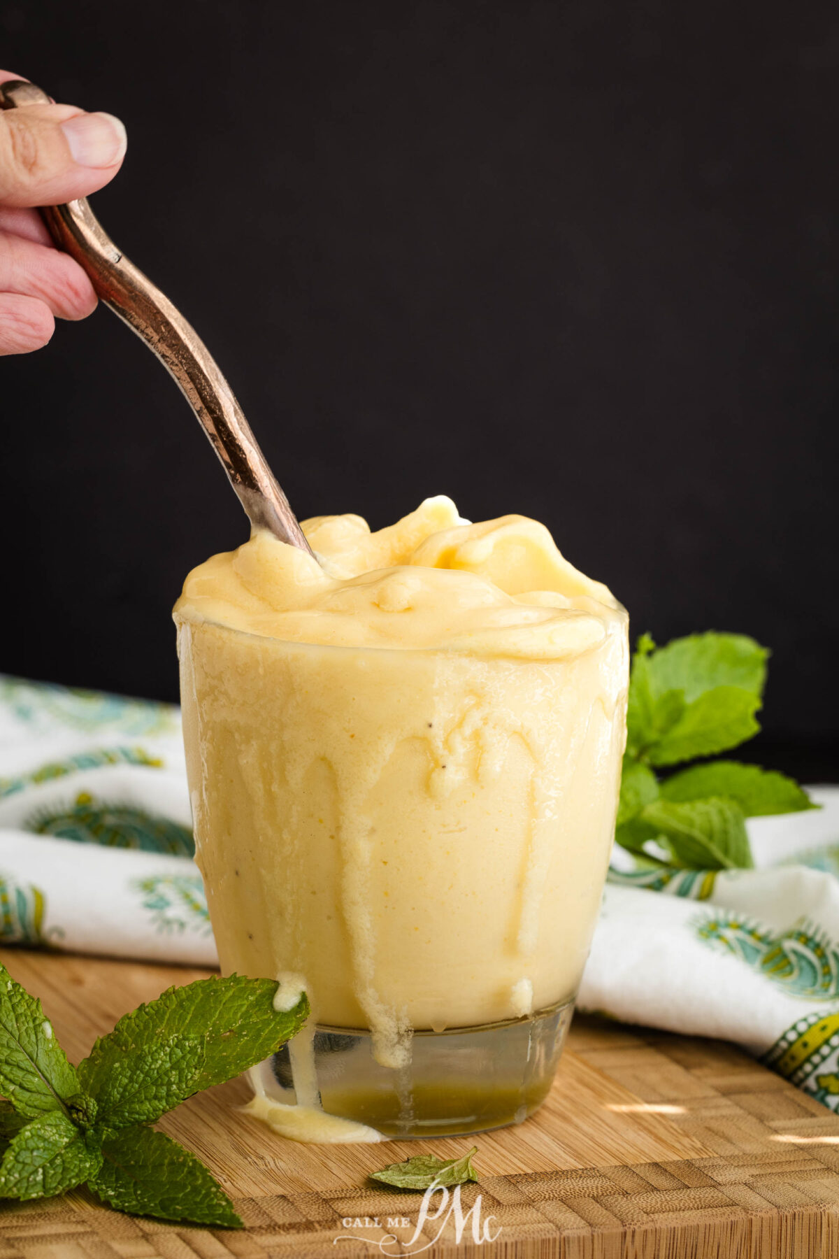A hand holds a spoon in a glass overflowing with Pineapple Coconut Smoothie, with some drips down the side. Fresh mint leaves and a patterned cloth decorate the wooden surface nearby.