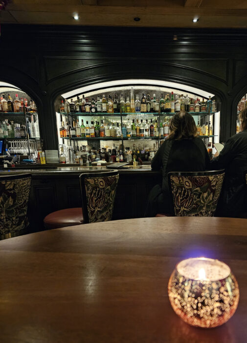 A dimly lit bar with shelves of liquor bottles, two people seated at the counter, and a decorative candle glowing on a wooden table in the foreground.