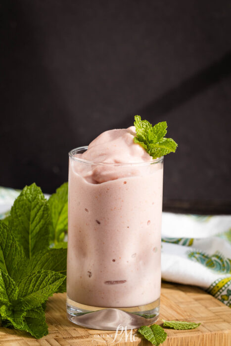 A glass of creamy pink smoothie garnished with a fresh mint sprig, placed on a wooden surface with mint leaves and a patterned cloth in the background.