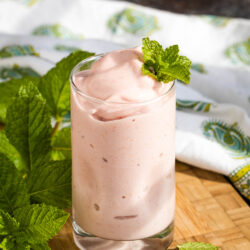 A glass of creamy pink smoothie garnished with a fresh mint sprig sits on a wooden board, with mint leaves and a patterned cloth in the background.