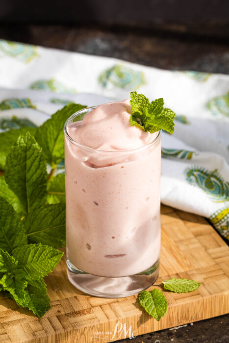 A glass of Strawberry Electrolyte Smoothie in a glass garnished with a fresh mint sprig sits on a wooden board, with mint leaves and a patterned cloth in the background.