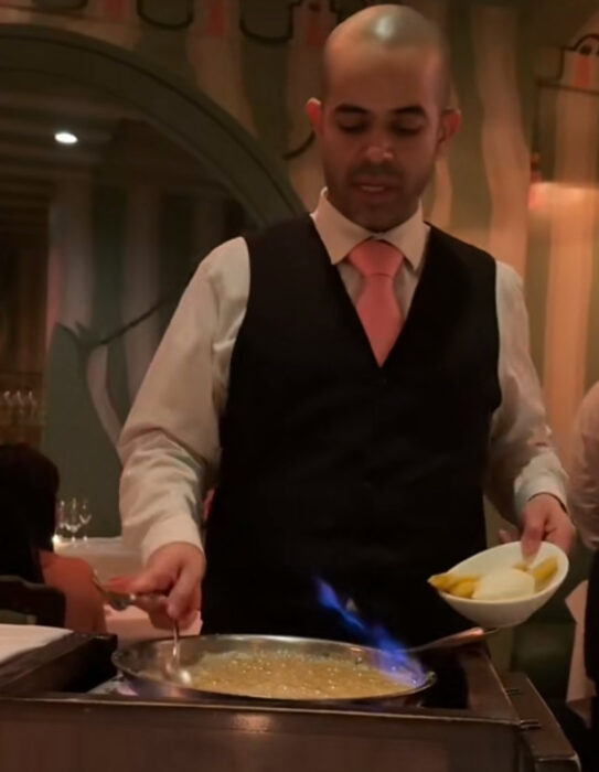 A server in a vest and tie prepares a flaming dish at a restaurant table, holding a bowl of ingredients and cooking with a blue flame in front of seated diners.