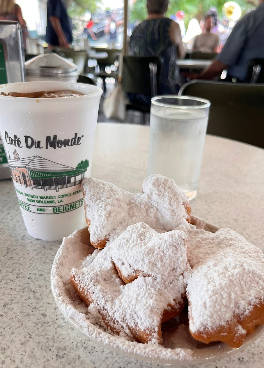 A plate of beignets covered in powdered sugar and a cup with “Café Du Monde” on it sit on a table, with a glass of water and blurred people in the background.