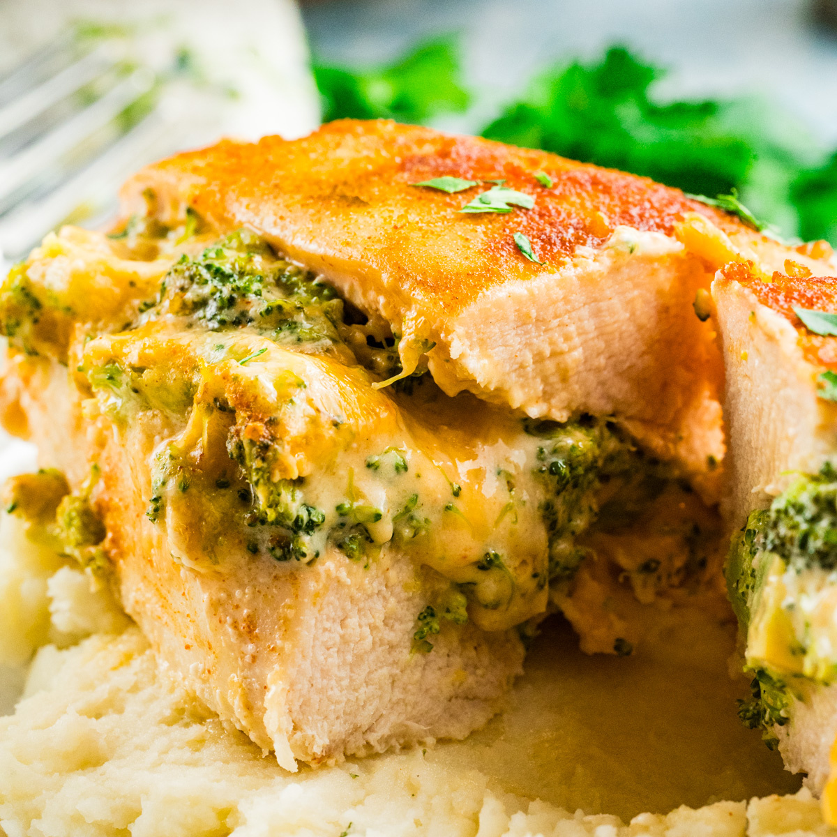 A close-up of sliced stuffed chicken breast filled with broccoli and melted cheese, served on a bed of creamy mashed potatoes, garnished with fresh herbs.