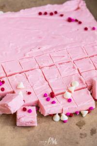 A slab of 2 Ingredient Pink Fudge is cut into small squares, with some pieces arranged in the foreground. The fudge is decorated with white chocolate chips and red, pink, and purple round sprinkles.