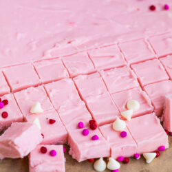 A slab of 2 Ingredient Pink Fudge is cut into small squares, with some pieces arranged in the foreground. The fudge is decorated with white chocolate chips and red, pink, and purple round sprinkles.