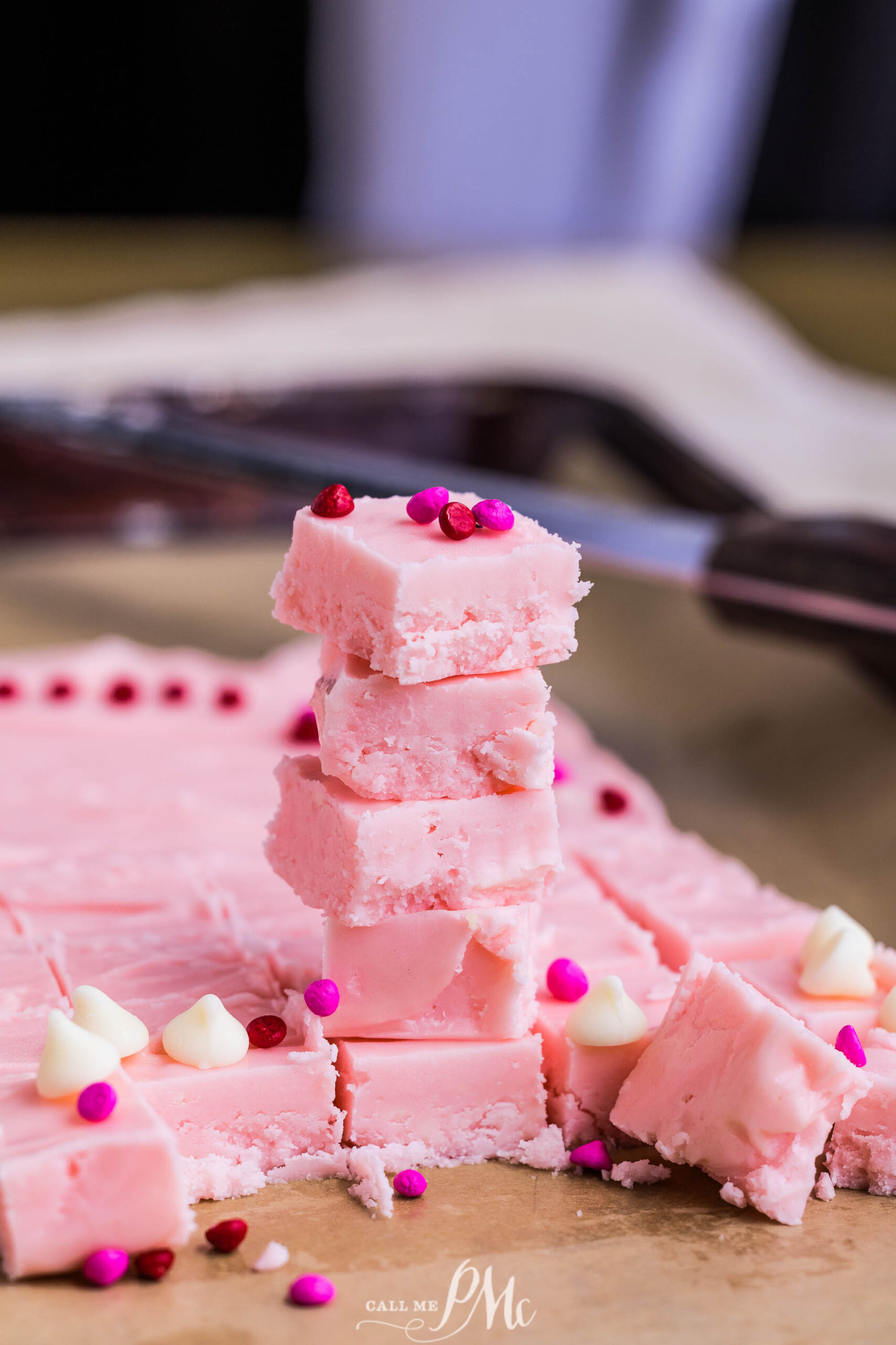 A stack of 2 Ingredient Pink Fudge squares topped with red and pink sprinkles, surrounded by more fudge pieces, white chocolate chips, and sprinkles on a wooden surface. A knife is visible in the background.
