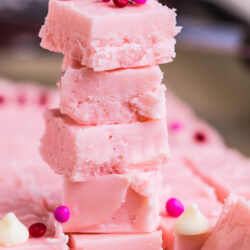 A stack of 2 Ingredient Pink Fudge squares with a smooth texture, topped with pink and red candy sprinkles, with more fudge pieces and white chocolate chips scattered in the background.