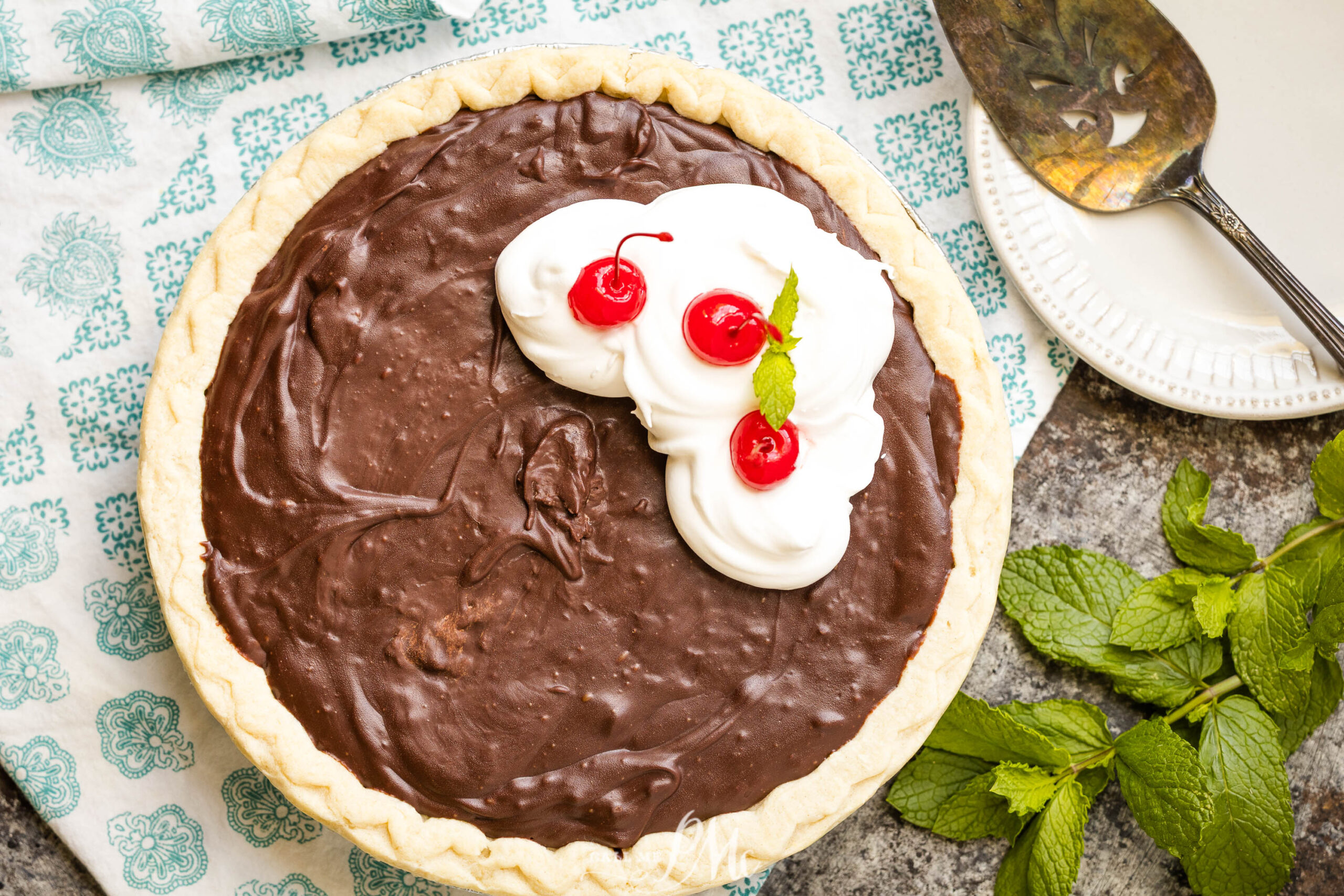 Slice of old-fashioned Coca-Cola Pie topped with whipped cream, three maraschino cherries, and mint leaves, sits on a patterned cloth with a serving spatula, plate, and fresh mint nearby.