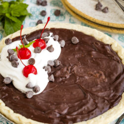 A chocolate pie with a flaky crust, topped with whipped cream, maraschino cherries, and chocolate chips, sits on a blue-patterned cloth next to plates, forks, and a sprig of fresh mint.