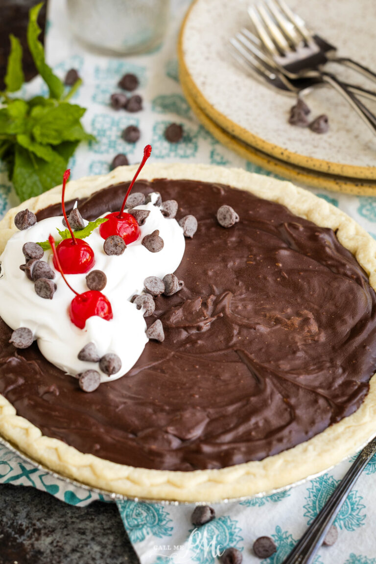 A chocolate pie with a flaky crust, topped with whipped cream, maraschino cherries, and chocolate chips, sits on a blue-patterned cloth next to plates, forks, and a sprig of fresh mint.