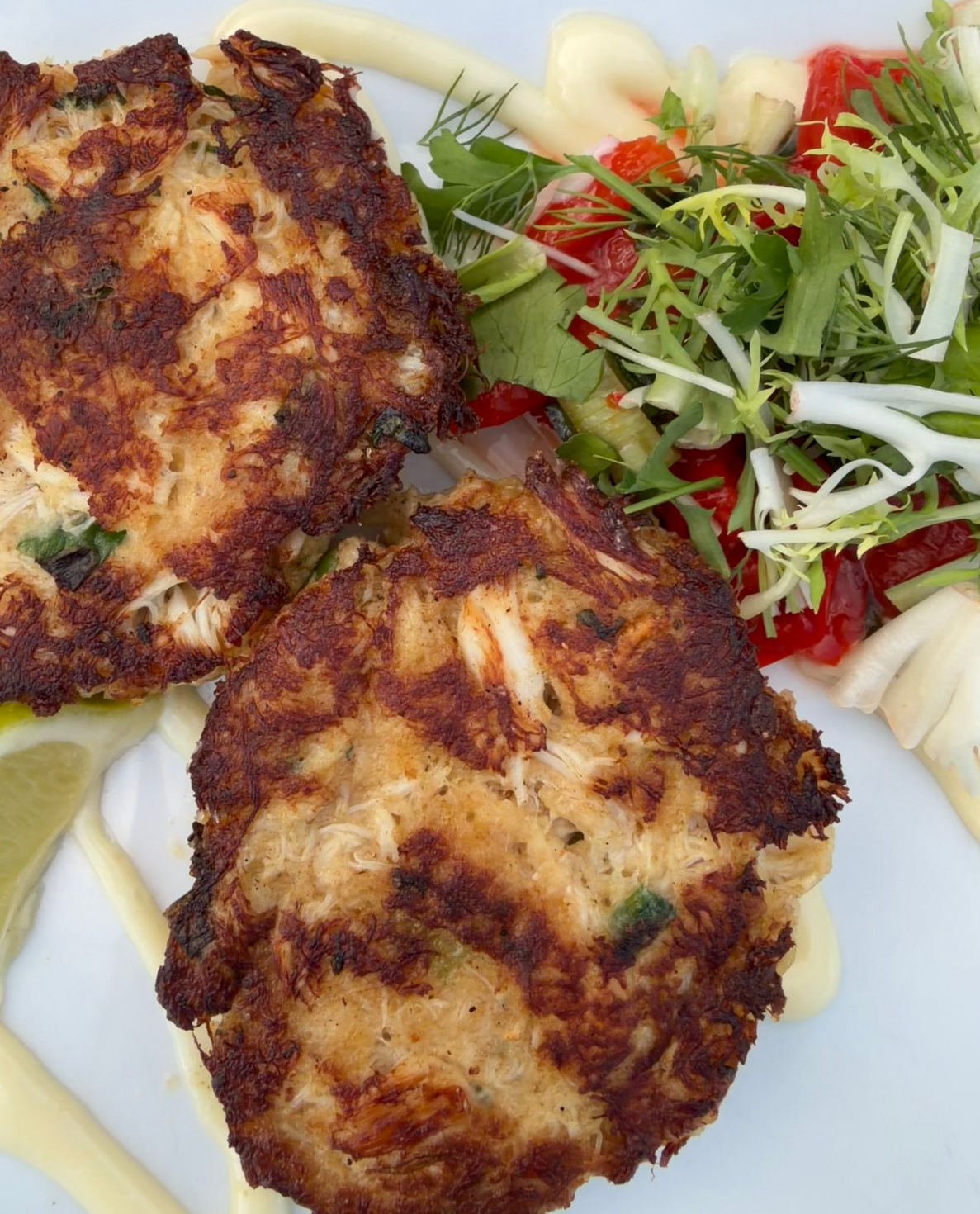 Two golden brown crab cakes on a white plate, garnished with fresh greens, red peppers, a lemon wedge, and drizzles of creamy sauce.