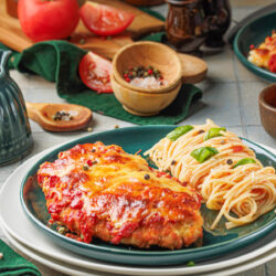 A plate with chicken parmesan, made using a classic Chicken Parmesan recipe, is topped with melted cheese and tomato sauce next to a serving of spaghetti garnished with basil. Fresh tomatoes, seasonings, and pepper grinders are in the background.