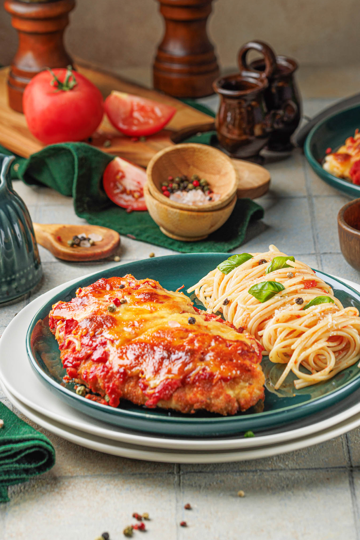 A plate with dinner is topped with melted cheese and tomato sauce next to a serving of spaghetti garnished with basil. Fresh tomatoes, seasonings, and pepper grinders are in the background.