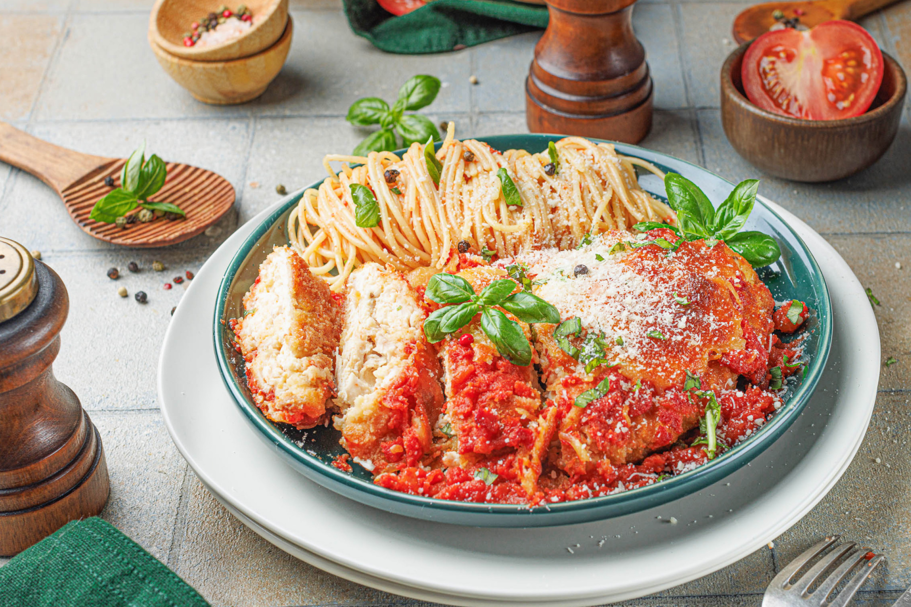 A classic Chicken Parmesan recipe comes to life on a plate of spaghetti with tomato sauce and breaded chicken cutlets, garnished with fresh basil and grated cheese. The dish sits on a table alongside pepper mills, tomato slices, and a wooden spoon, inviting you to enjoy this Italian favorite.