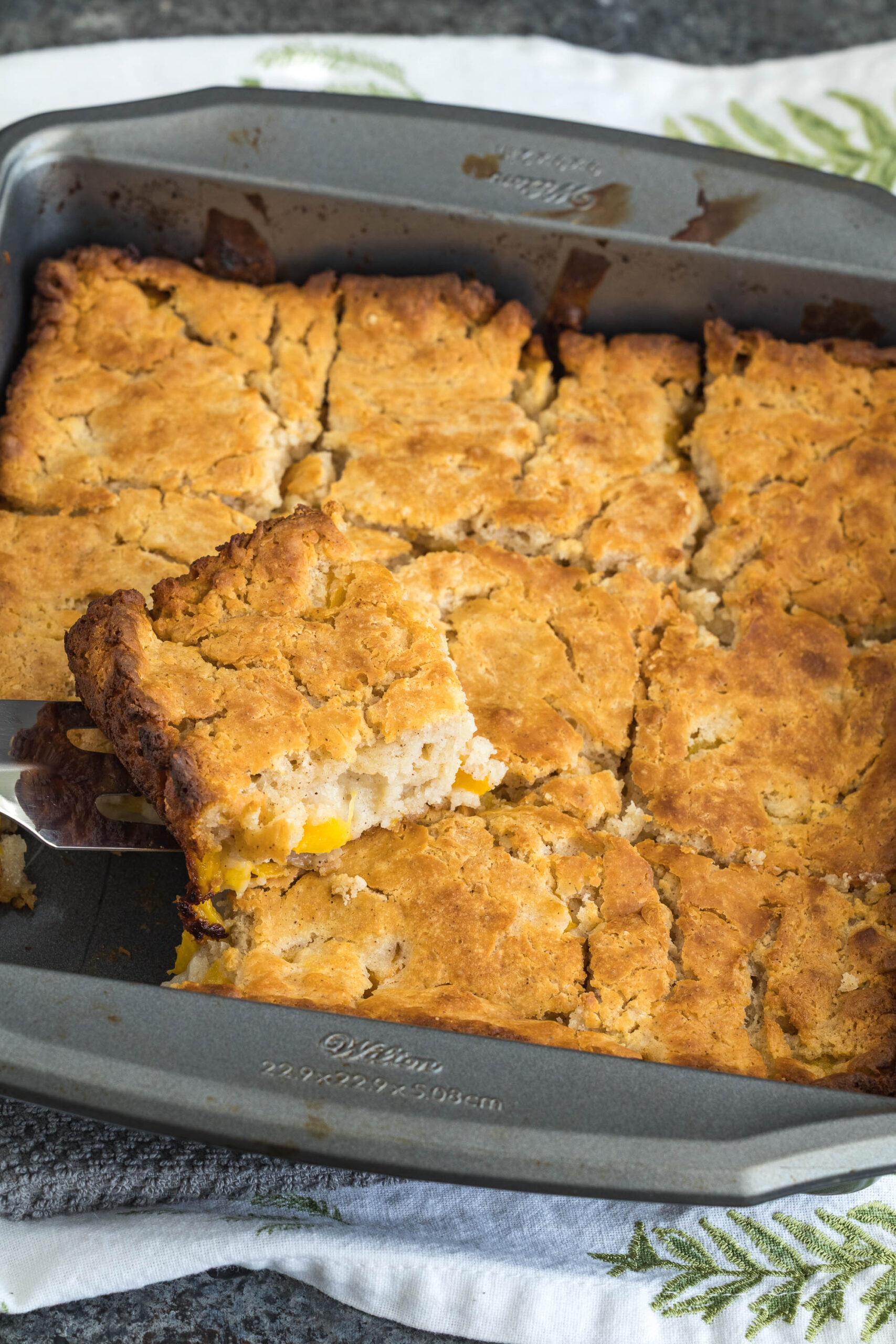 Golden-brown bread with a piece being lifted from a metal baking pan, revealing a filling that appears to include chunks of fruit. The pan rests on a patterned cloth.