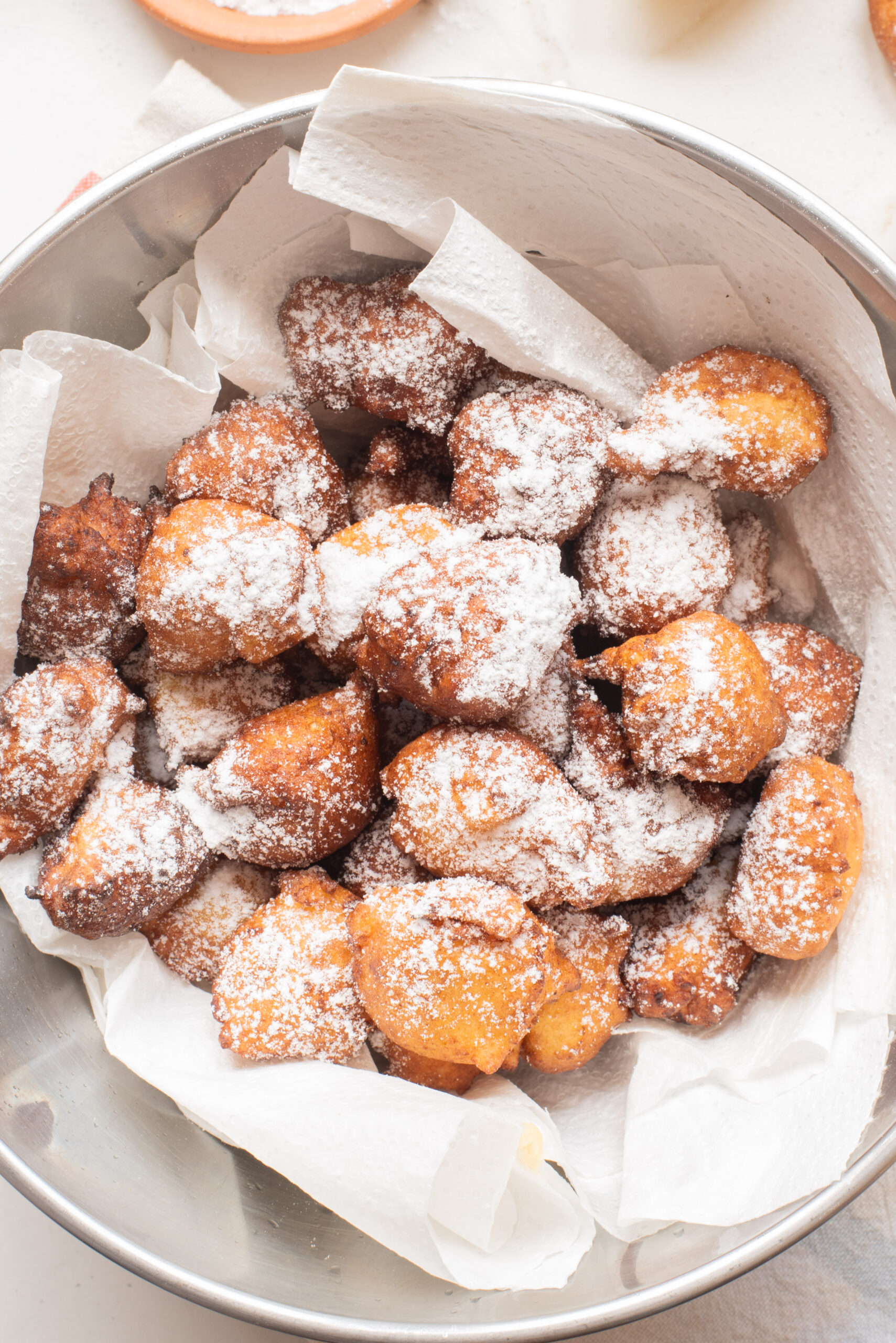 A metal bowl lined with paper towels holds golden brown ricotta fritters dusted with powdered sugar.