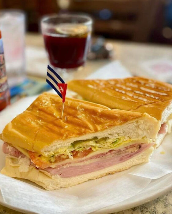 A Cuban sandwich cut in half, filled with ham, pork, cheese, pickles, and mustard, sits on white paper—a must-try for anyone wondering where to eat in Key West. A small Cuban flag toothpick is inserted in the sandwich, while a drink in a glass is blurred in the background.