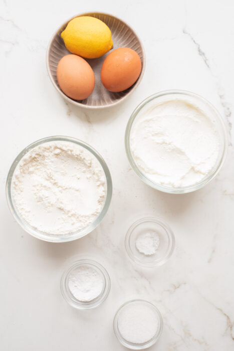 Two brown eggs and a lemon in a small bowl, with bowls of flour, sugar, and soft cheese.