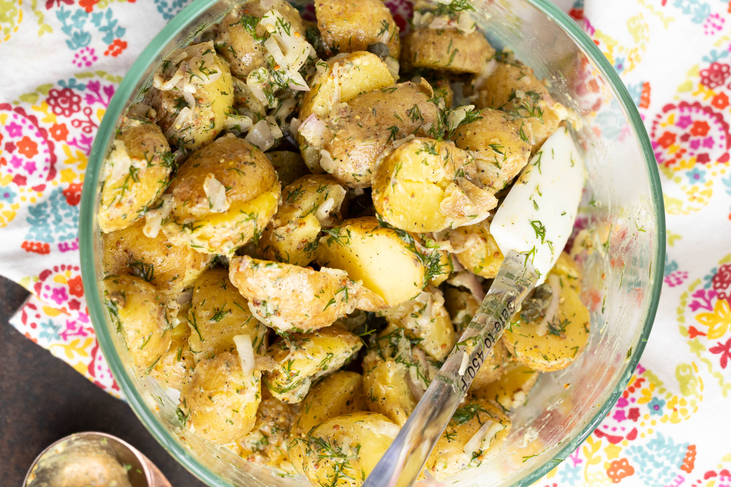 A glass bowl filled with sliced yellow potatoes mixed with herbs and onions, resting on a colorful floral-patterned cloth. Perfectly embodying Classic French Potato Salad, a serving spoon is in the bowl.