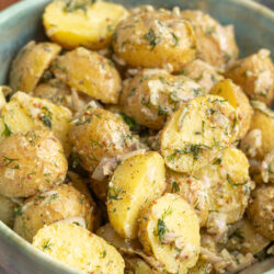 A bowl of Classic French Potato Salad with halved yellow potatoes, herbs, and chopped onions, served in a green ceramic bowl on a gray surface.