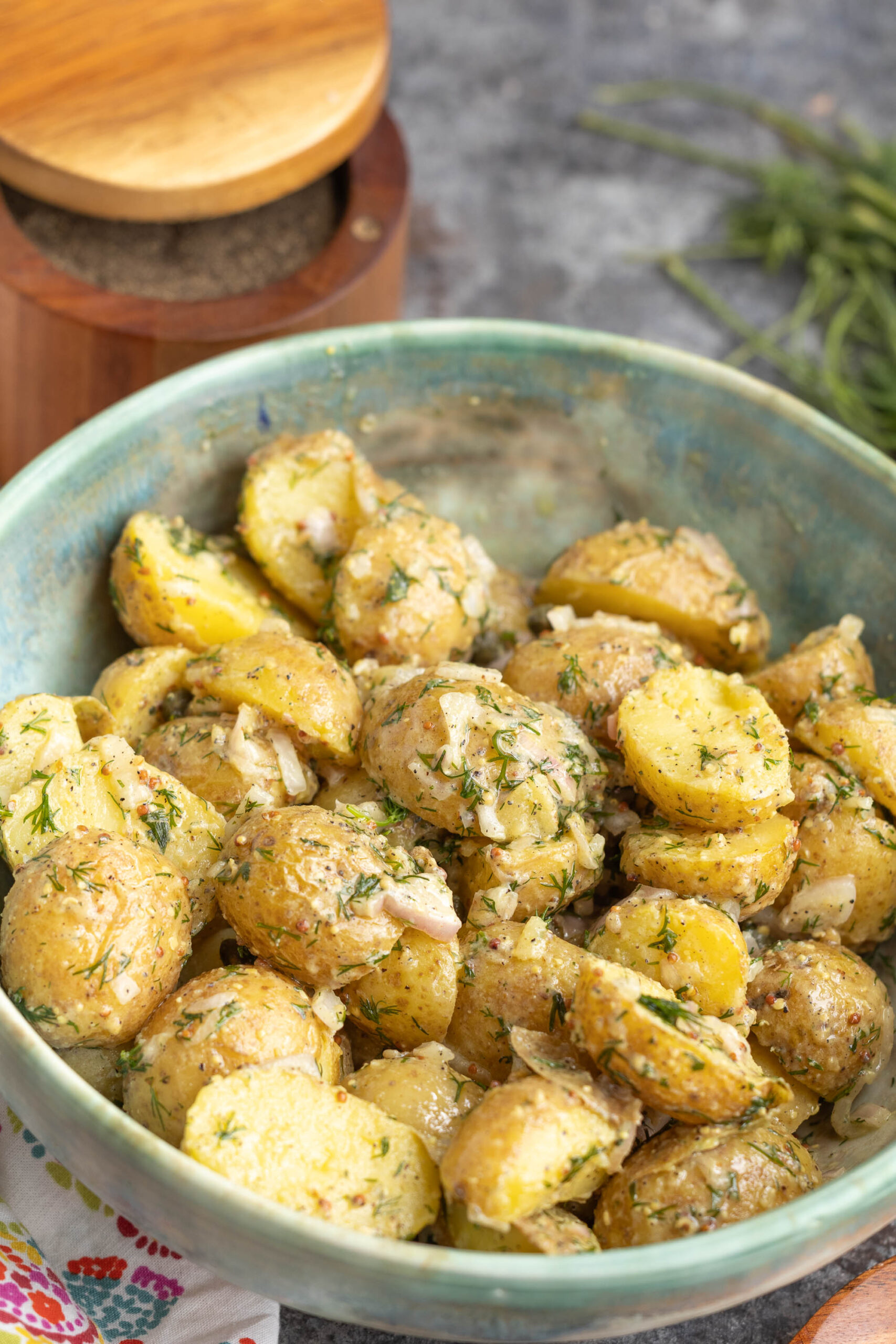 A ceramic bowl filled with sliced baby vegetables coated in herbs and dressing, garnished with fresh dill, sits on a table beside a wooden pepper grinder.
