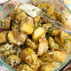 A glass bowl filled with creamy Classic French Potato Salad, garnished with chopped dill and onions, with a spoon resting inside. The bowl sits on a colorful, floral-patterned cloth.