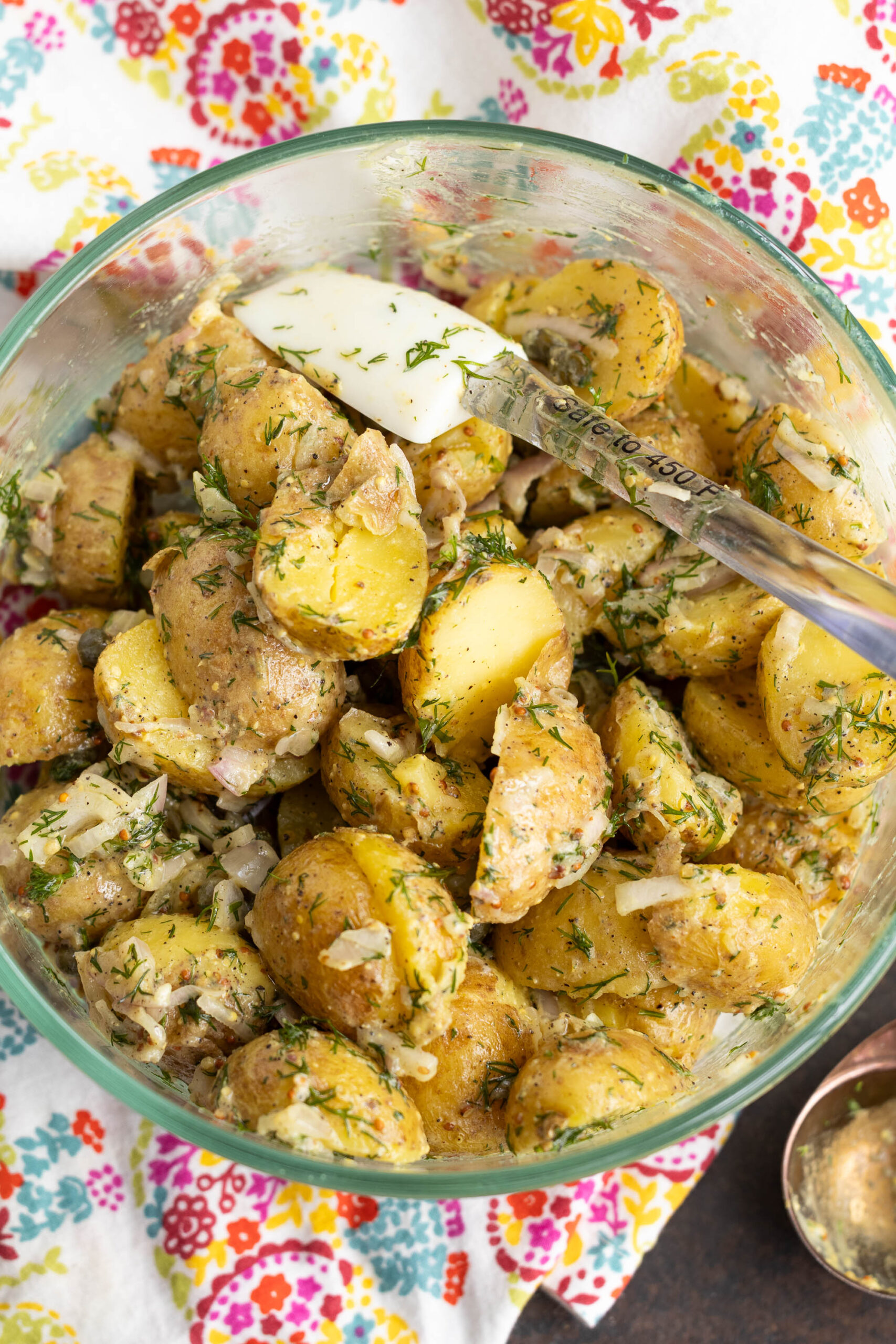 A glass bowl filled with creamy Classic French Potato Salad, garnished with chopped dill and onions, with a spoon resting inside. The bowl sits on a colorful, floral-patterned cloth.