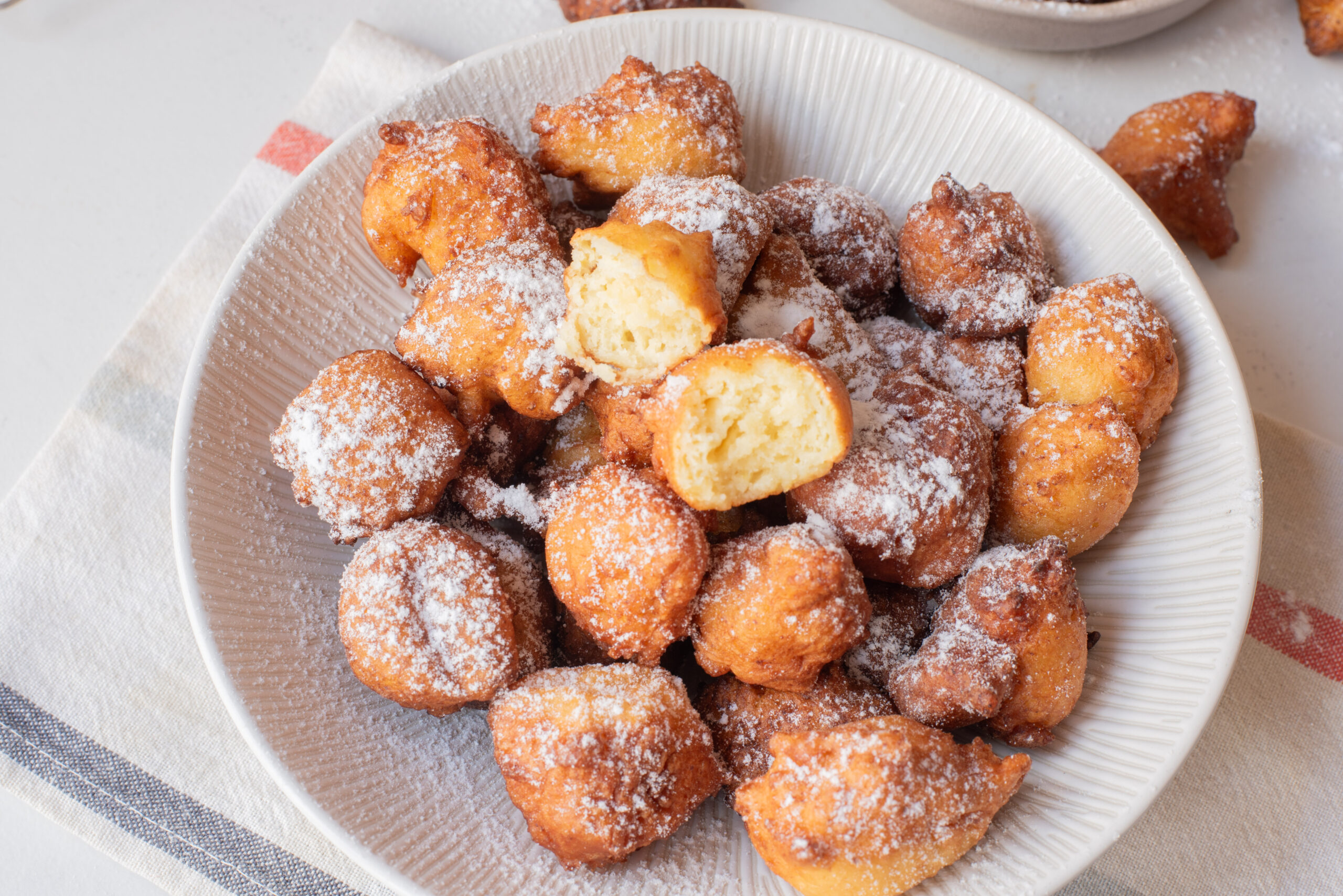A white bowl filled with fried bread nuggets dusted with powdered sugar, with one fritter broken open to show the soft, fluffy inside. The bowl sits on a striped cloth.