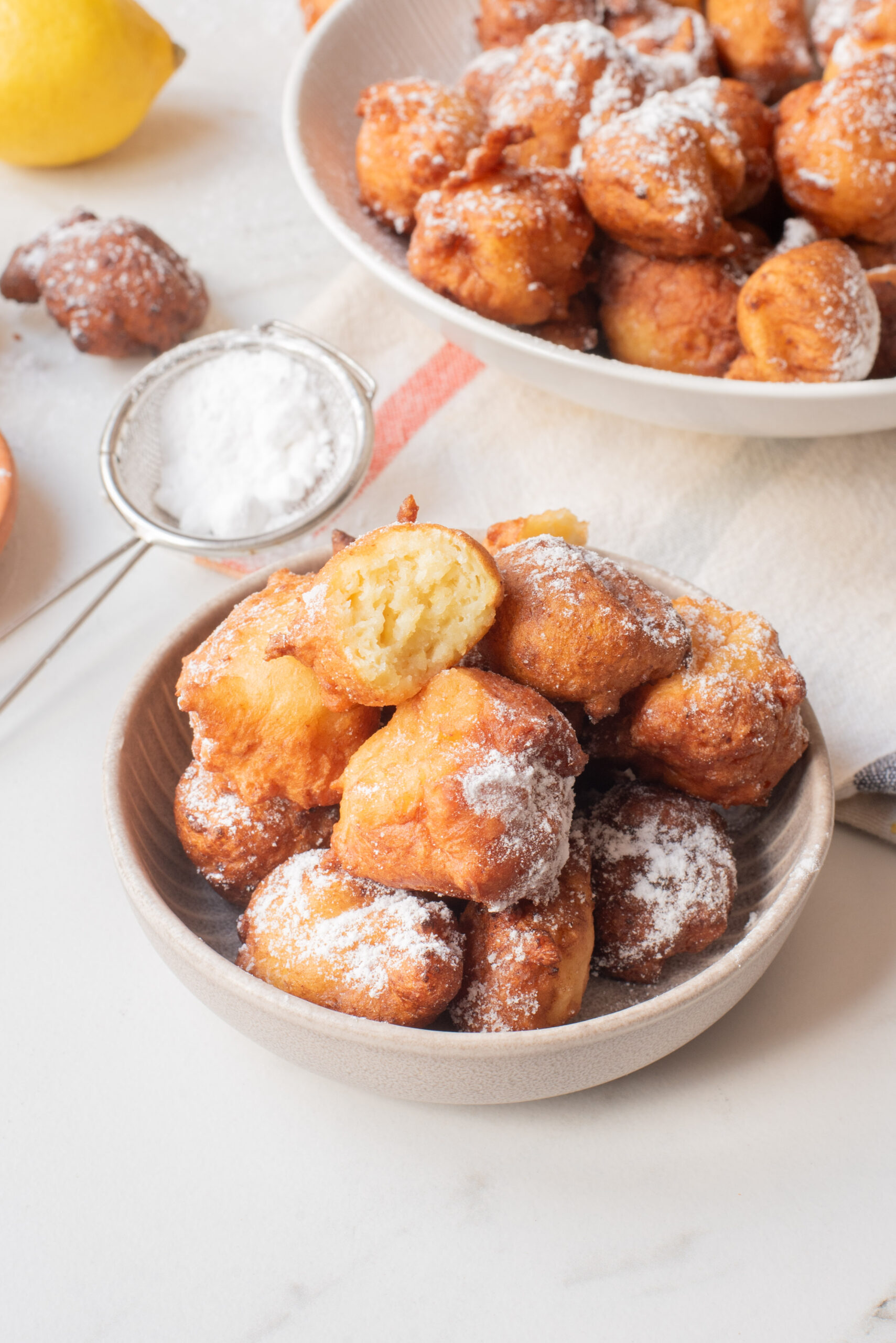 A bowl of golden, round fritters dusted with powdered sugar sits on a white surface, with one fritter broken open to show its soft interior. A plate of more fritters and a sifter of powdered sugar are in the background.