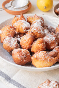 A plate of golden-brown fritters dusted with powdered sugar, with a lemon and a sifter in the background on a light-colored tablecloth.