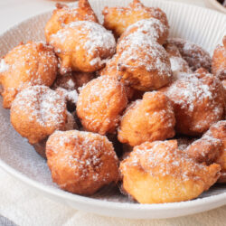 A plate of golden-brown fritters dusted with powdered sugar, with a lemon and a sifter in the background on a light-colored tablecloth.