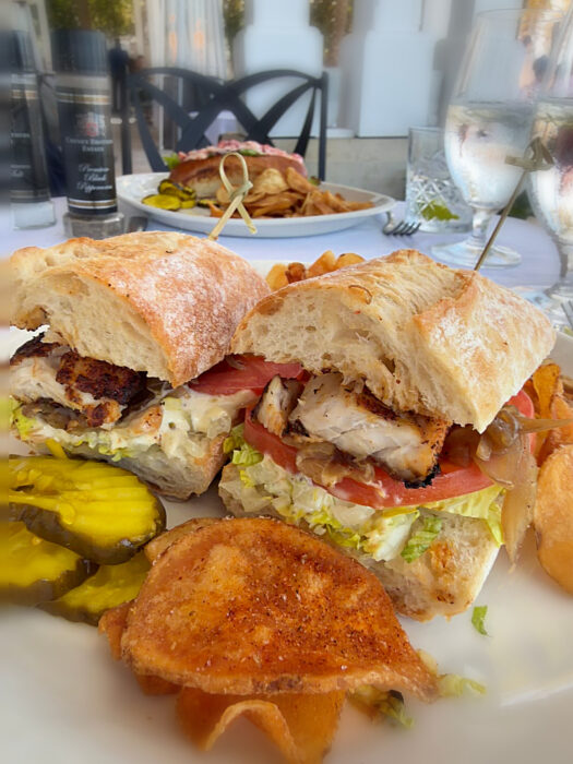 A close-up of a grilled chicken sandwich with lettuce, tomato, and pickles on ciabatta bread, served with seasoned chips and water glasses on a dining table outdoors—perfect inspiration if you’re searching for where to eat in Key West.