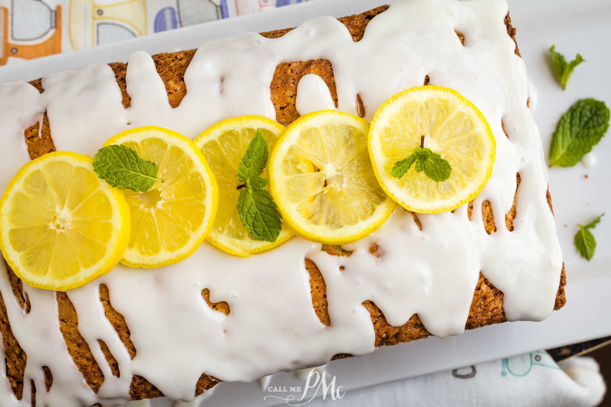 A loaf of Lemon Zucchini Bread topped with white icing, garnished with four lemon slices and two sprigs of fresh mint, displayed on a white tray.