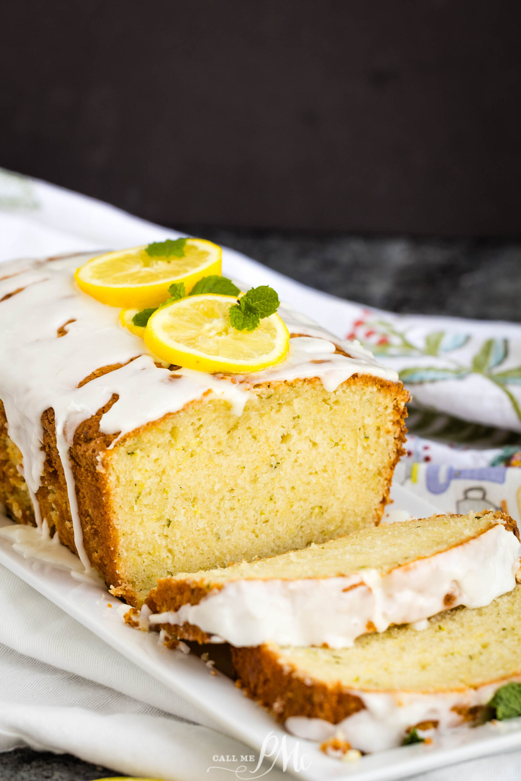 A loaf of glazed Lemon Zucchini Bread with two lemon slices and mint leaves on top, sitting on a white plate. One slice is cut and lies in front of the loaf.