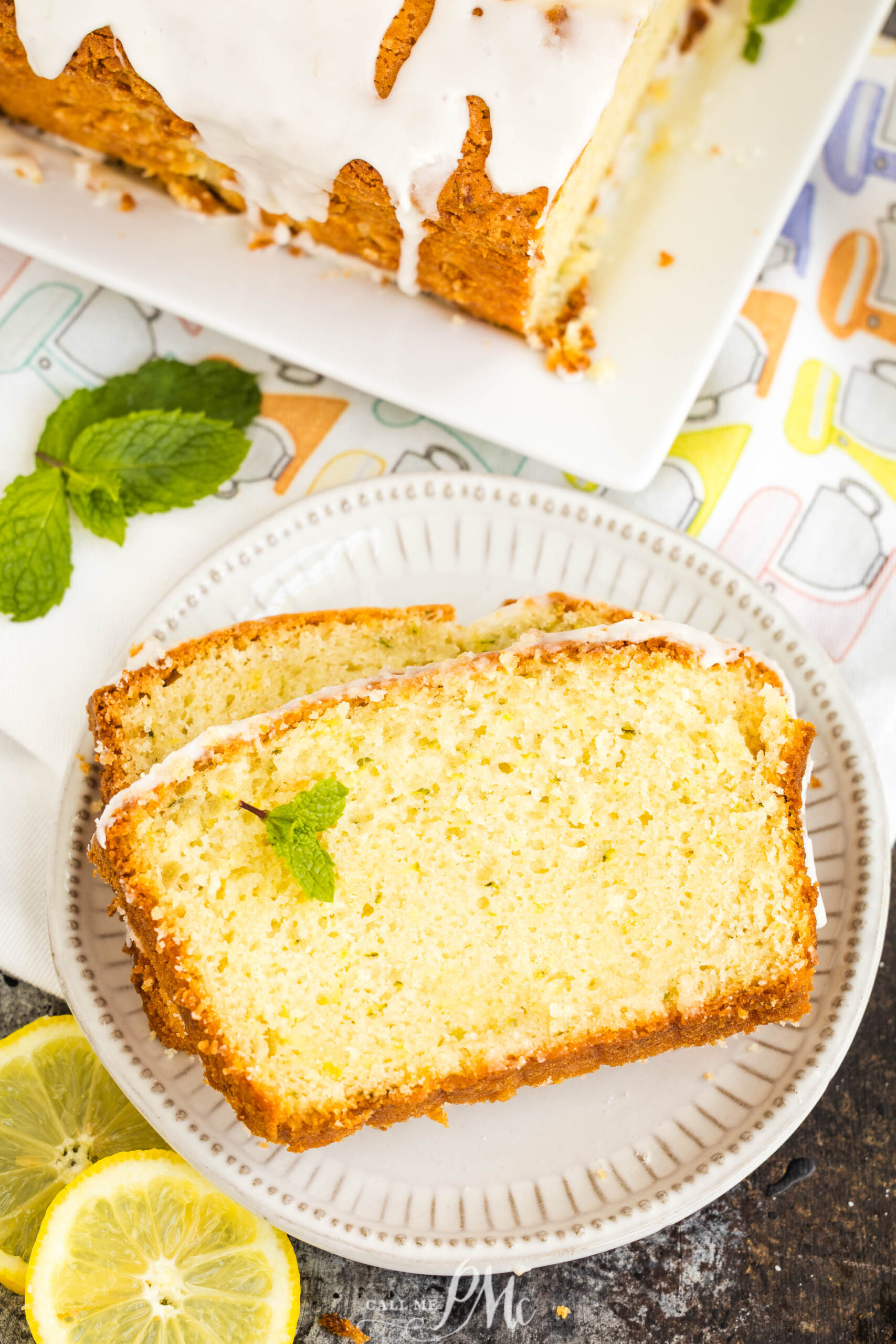 Two slices loaf cake are on a plate with lemon slices and mint, with more cake on a platter in the background.
