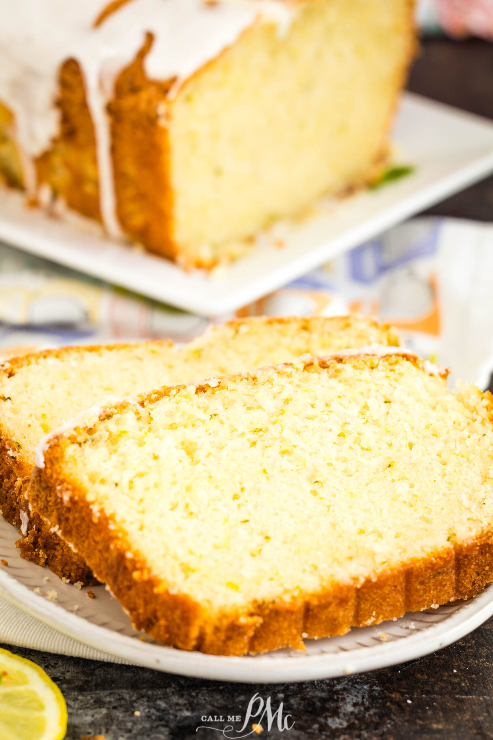 Two slices of lemon loaf cake with icing on a white plate, with the rest of the loaf and a lemon slice in the background—an irresistible scene reminiscent of freshly baked Lemon Zucchini Bread, perfect for brightening any day.