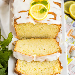 A loaf of Lemon Zucchini Bread with white icing, garnished with lemon slices and mint. Two slices are cut and laid in front on a white platter, accompanied by fresh mint and lemon slices nearby.