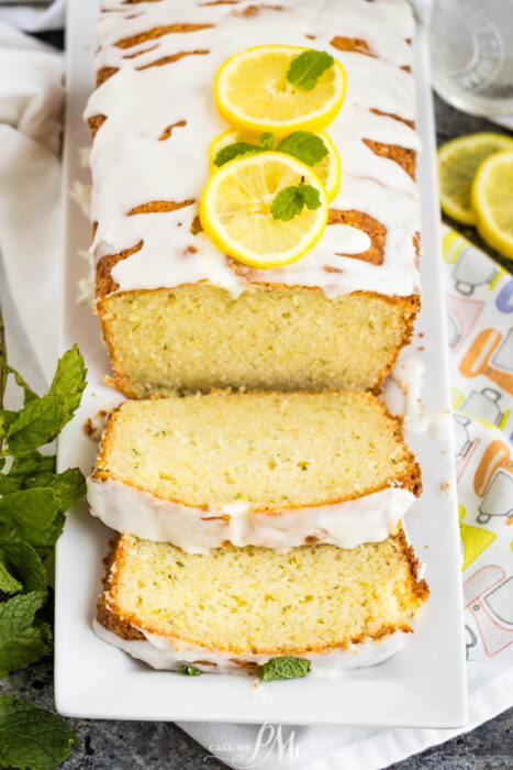 A loaf of Lemon Zucchini Bread with white icing, garnished with lemon slices and mint. Two slices are cut and laid in front on a white platter, accompanied by fresh mint and lemon slices nearby.