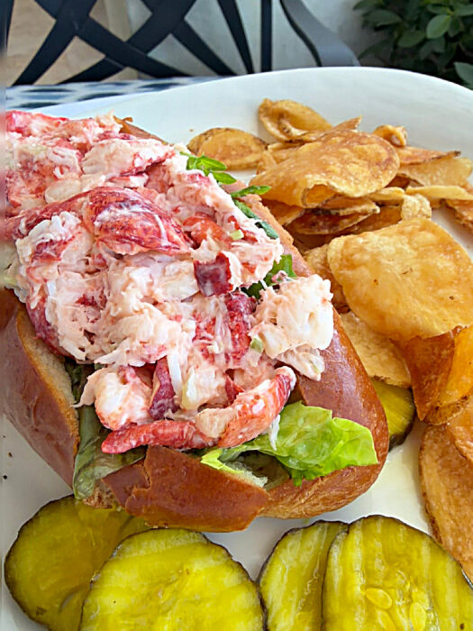 A lobster roll with leafy greens in a bun, served with potato chips and pickle slices on a white plate—a must-try for anyone searching for where to eat in Key West.