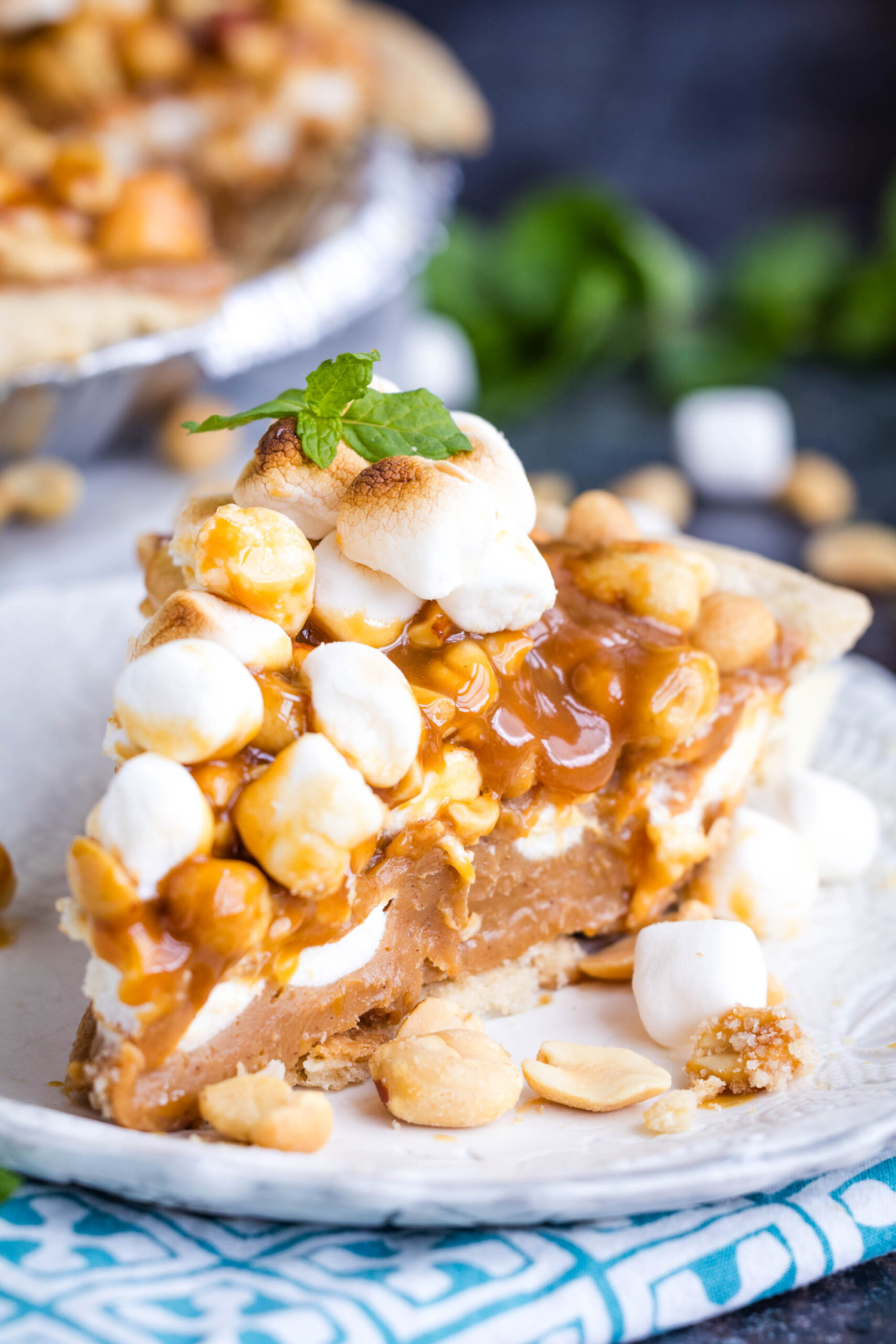 A slice of Payday Pie topped with caramel, marshmallows, and peanuts sits on a white plate, with more marshmallows and peanuts scattered around. A mint leaf garnishes the pie.