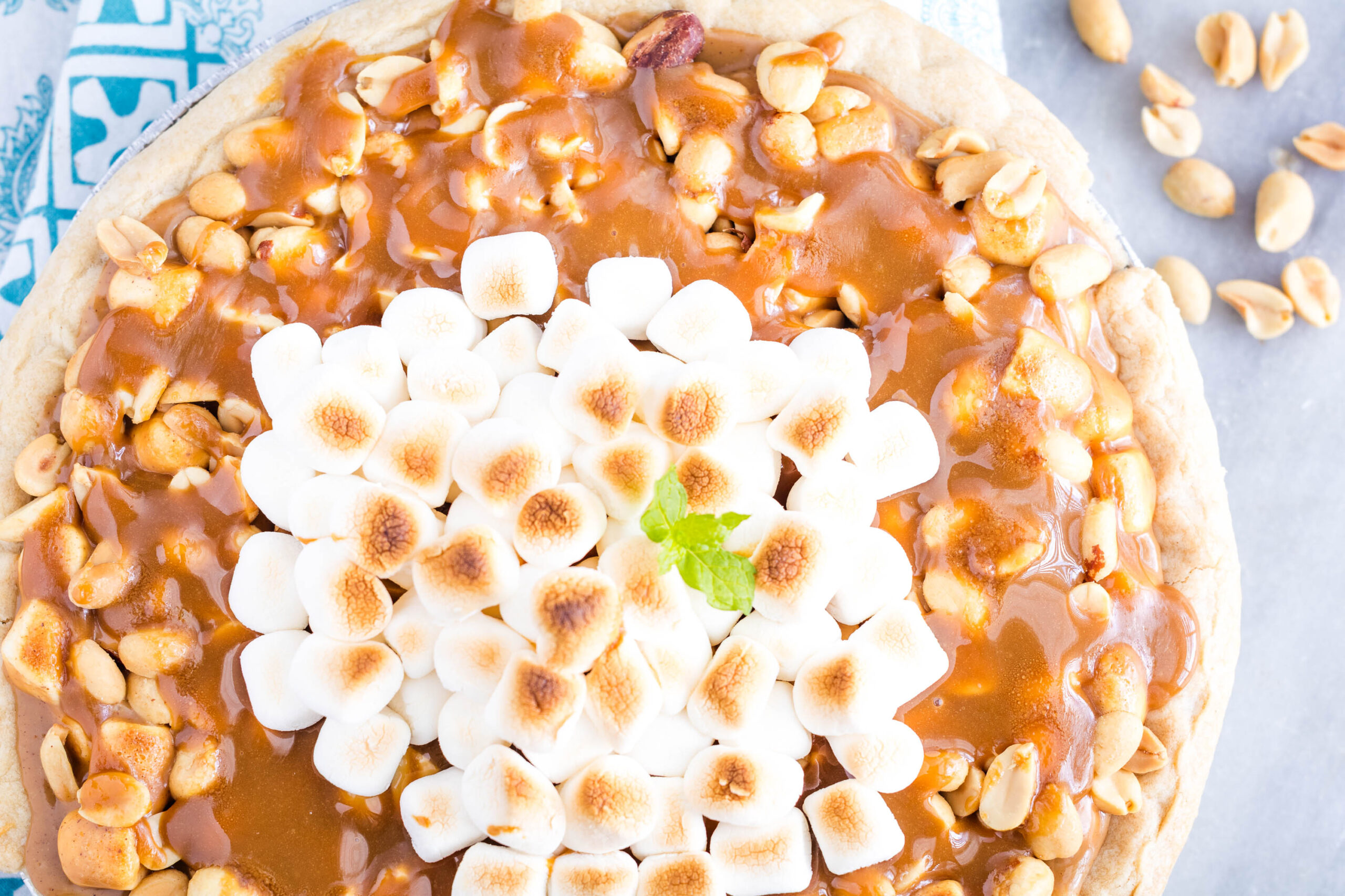 A Payday Pie topped with toasted marshmallows, caramel sauce, and peanuts sits on a blue-patterned cloth, with a few peanuts scattered on the side.