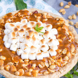 A decadent Payday Pie topped with caramel, peanuts, and toasted marshmallows, garnished with a sprig of mint. Scattered peanuts and mint leaves surround the pie on a marble surface.