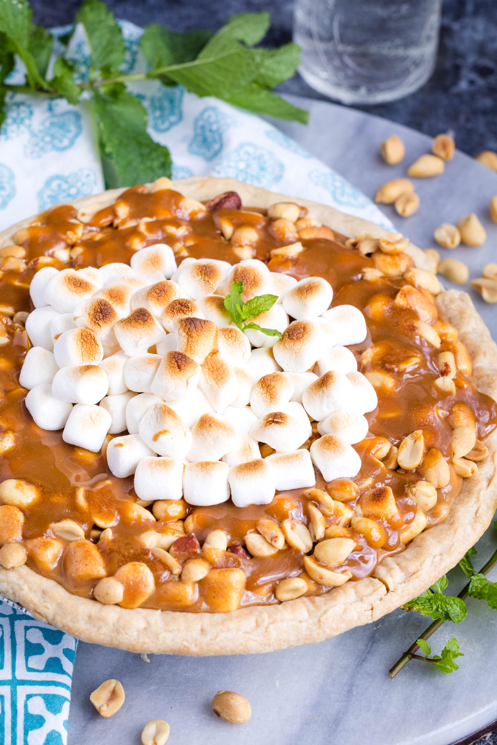 A decadent Payday Pie topped with caramel, peanuts, and toasted marshmallows, garnished with a sprig of mint. Scattered peanuts and mint leaves surround the pie on a marble surface.
