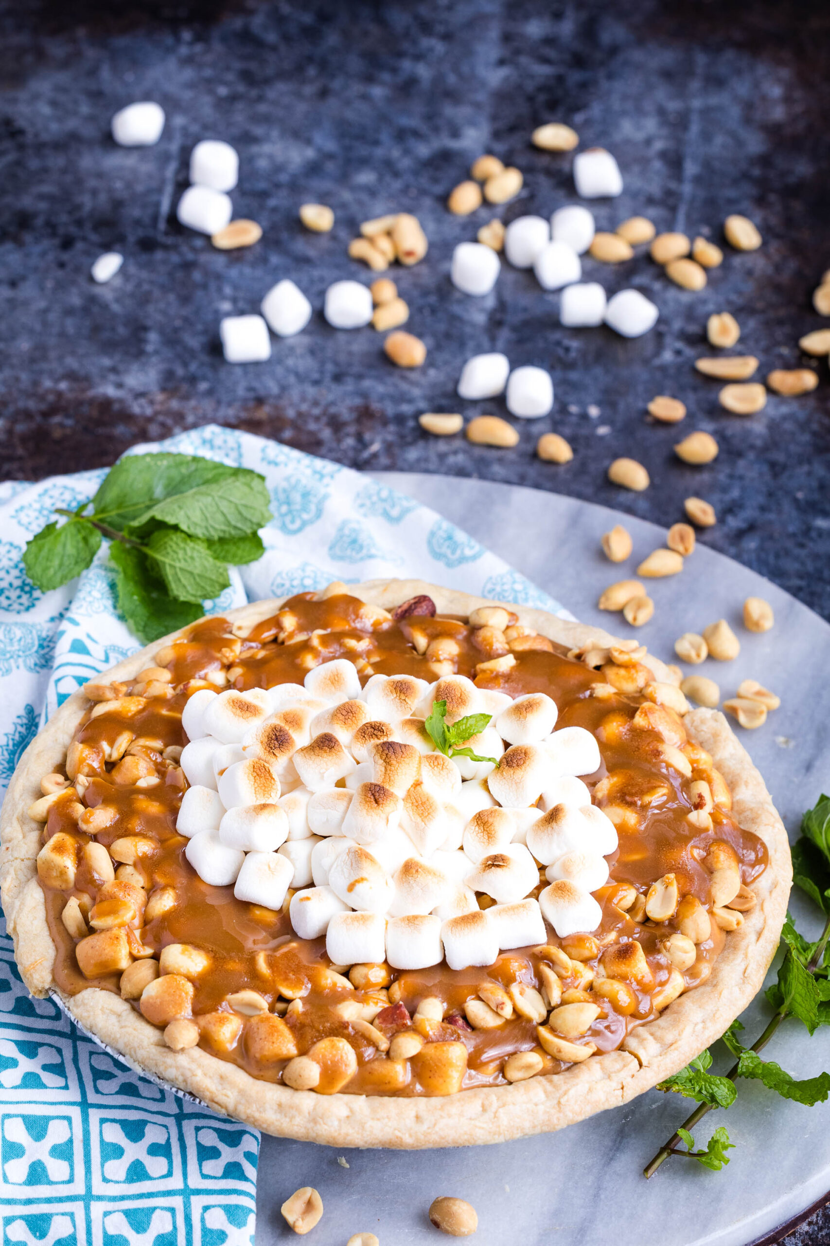 A decadent Payday Pie topped with toasted mini marshmallows and peanuts sits on a marble surface with a blue napkin, mint garnish, and scattered peanuts and marshmallows nearby.