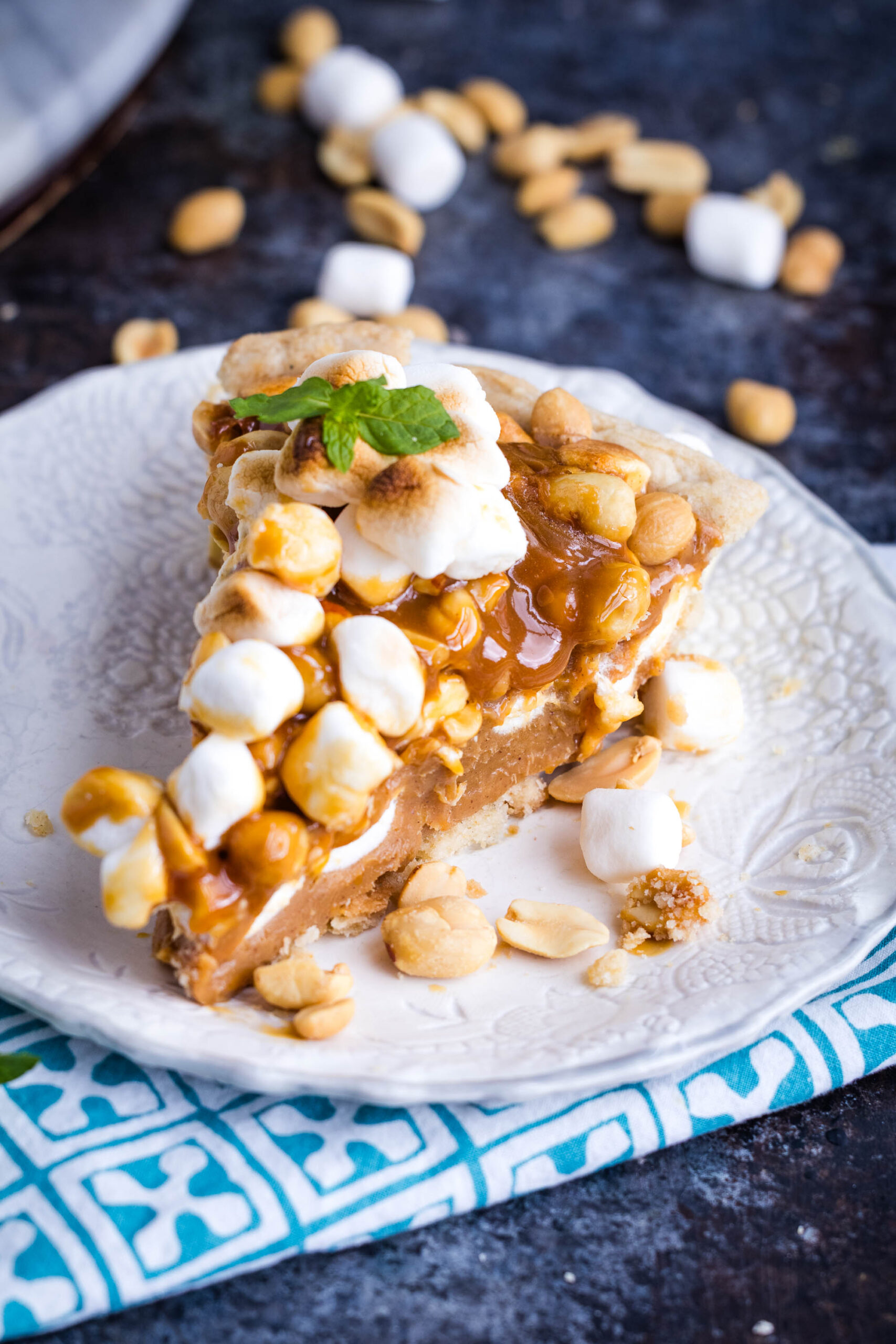 A decadent slice of Payday Pie topped with toasted marshmallows, peanuts, caramel sauce, and a mint leaf sits on a white textured plate with scattered peanuts and marshmallows around it.