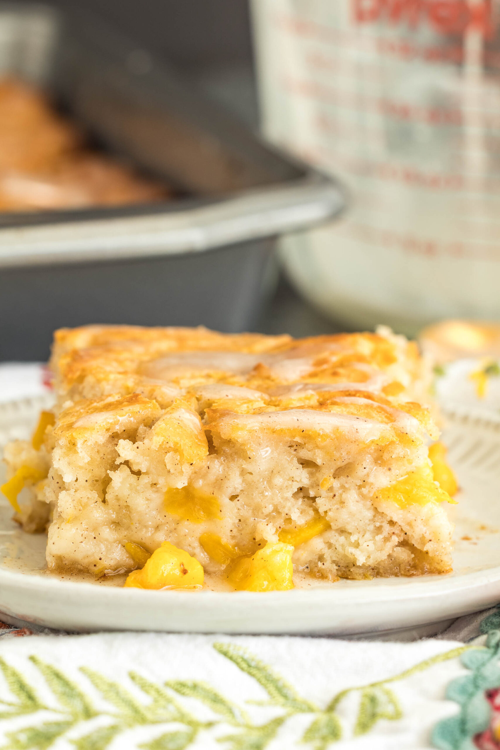 A close-up of a slice of bread, resting on a patterned plate. In the background, a baking pan and a measuring cup are blurred—setting the scene for a comforting kitchen moment.