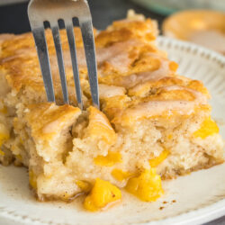 A close-up shot of a fork piercing a moist square of peach cobbler cake on a white plate. The cake is glazed, golden-brown, and filled with chunks of yellow peaches—evoking the irresistible flavors of Peach Butter Swim Biscuits. A corner of a baking pan is visible in the blurred background.