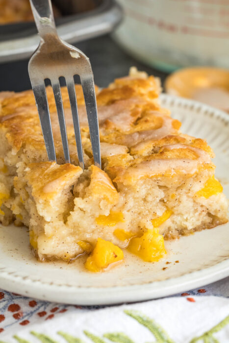 A close-up shot of a fork piercing a moist square of peach cobbler cake on a white plate. The cake is glazed, golden-brown, and filled with chunks of yellow peaches—evoking the irresistible flavors of Peach Butter Swim Biscuits. A corner of a baking pan is visible in the blurred background.