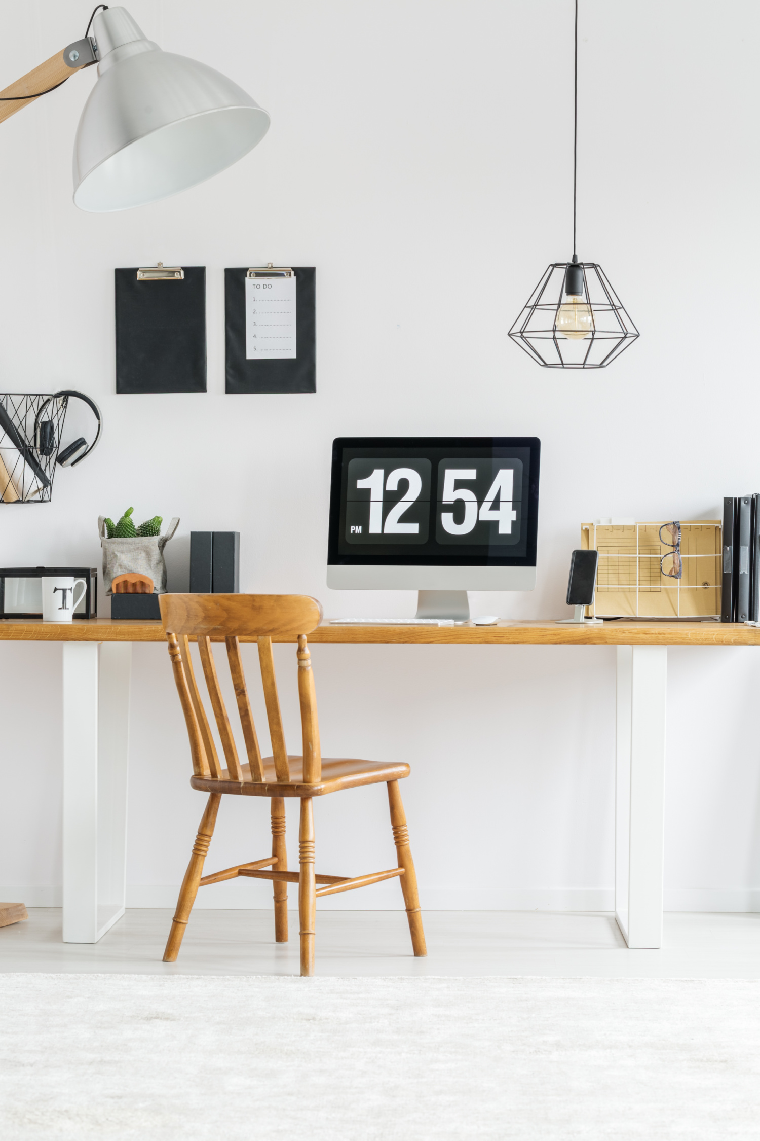 Minimalist workspace with a wooden chair, desk, computer displaying 12:54, lamp, hanging light, clipboards on the wall, and organized office supplies against a white background—an ideal setup for anyone interested in how to successfully work from home.
